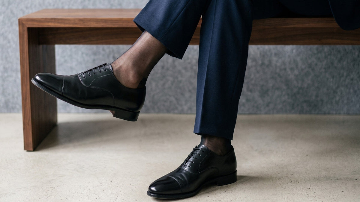 Man in a navy suit seated on a walnut bench, legs crossed, wearing black sheer dress socks and black cap toe oxfords