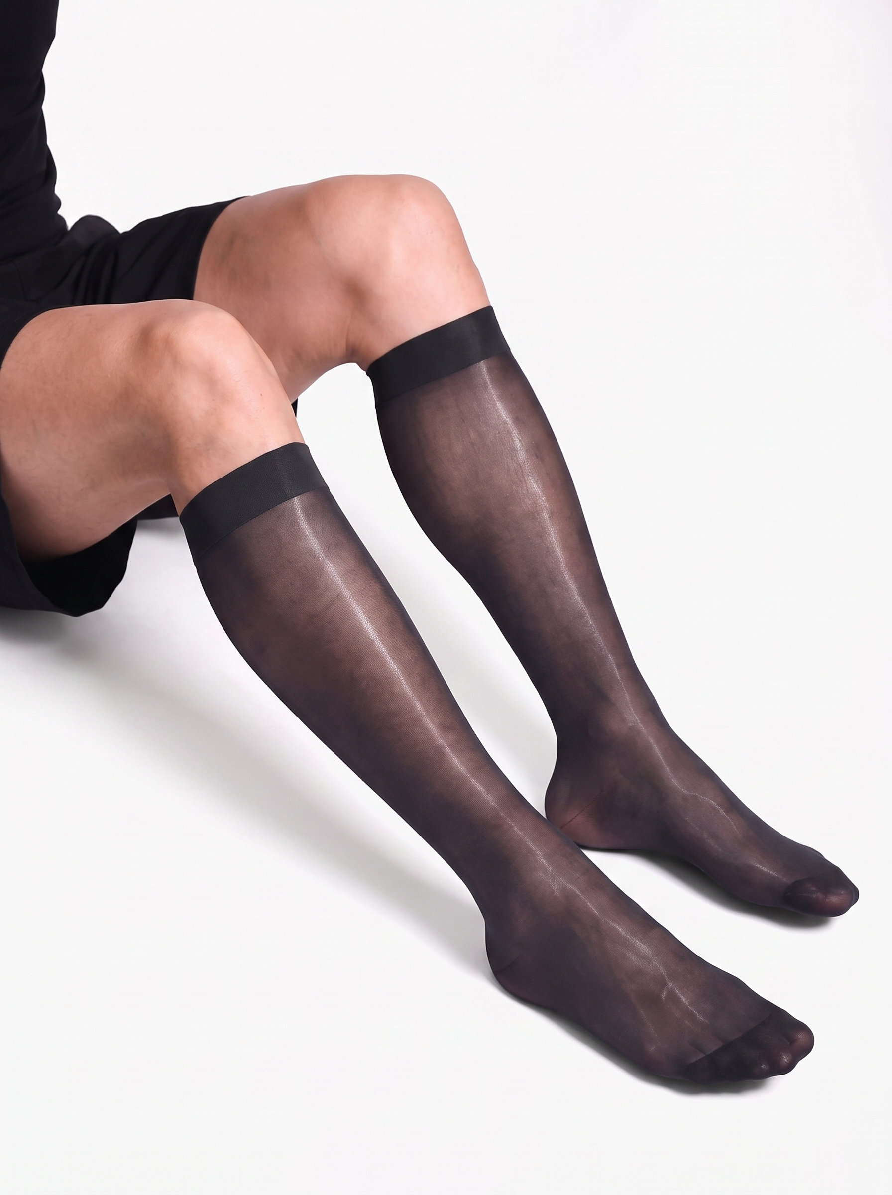 Man seated with knees bent wearing NYLO 10D sheer black dress socks below the knee with solid cuffs