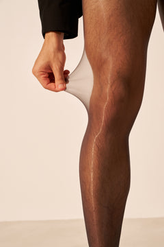 A close-up of a person's leg wearing sheer black tights, with a hand pulling the fabric upward to demonstrate stretch and transparency against a plain light beige studio background.