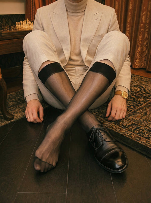Person sitting on a wooden floor wearing a beige suit, black shoes, and black tights.