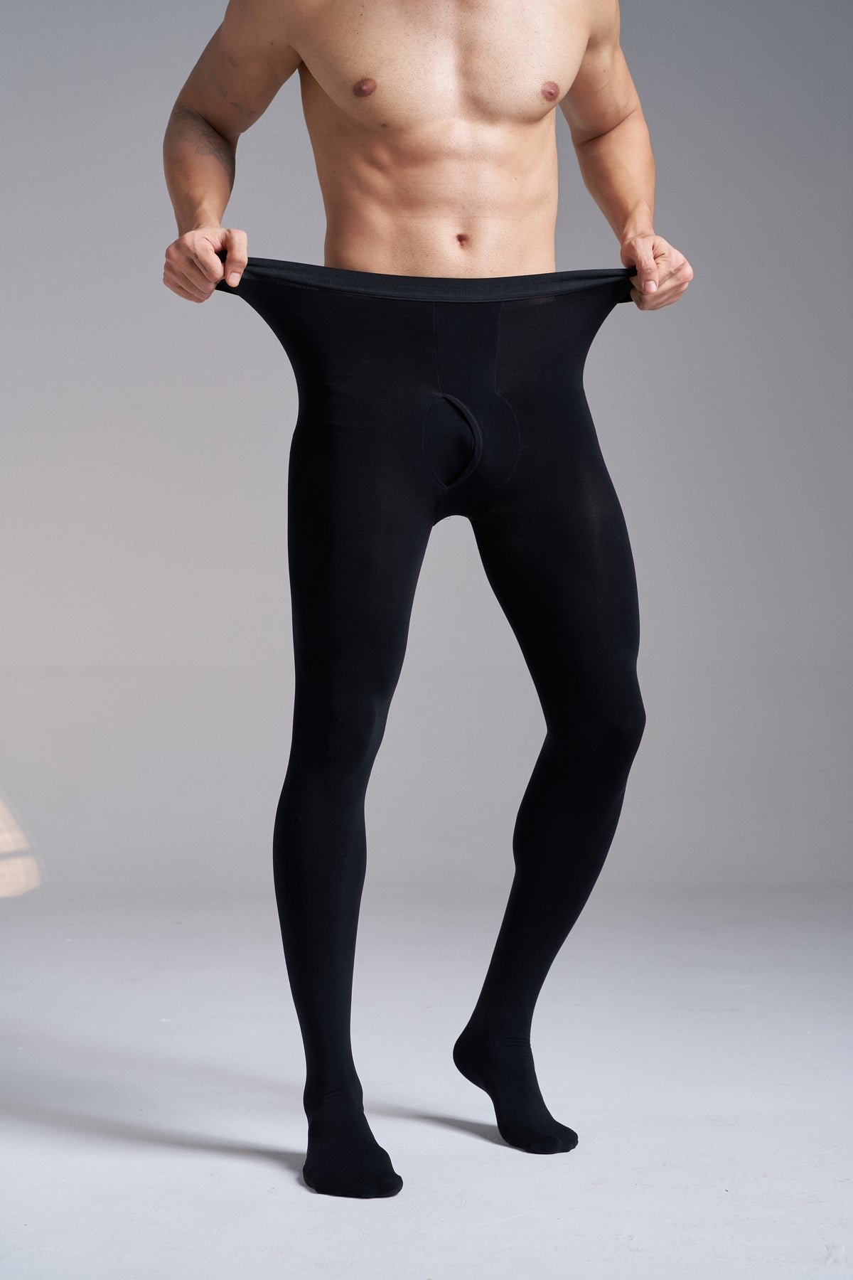 Front view of a man wearing NYLO 120D Opaque Tights in black, pulling the knitted waistband out at both sides, with the underwear-inspired front visible.