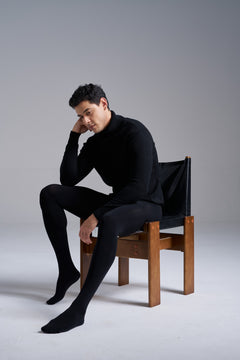 Man in a black turtleneck seated on a wooden chair, wearing NYLO 120D Opaque Tights in black.