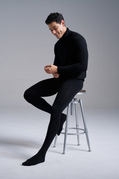 Man in a black turtleneck seated on a metal stool, wearing NYLO 120D Opaque Tights in black.