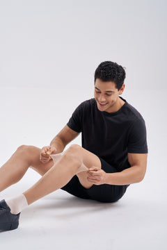 Man in a black t-shirt and shorts seated on the floor, wearing NYLO 10D Sock Foot Sheer Tights with white sock feet and black sneakers.