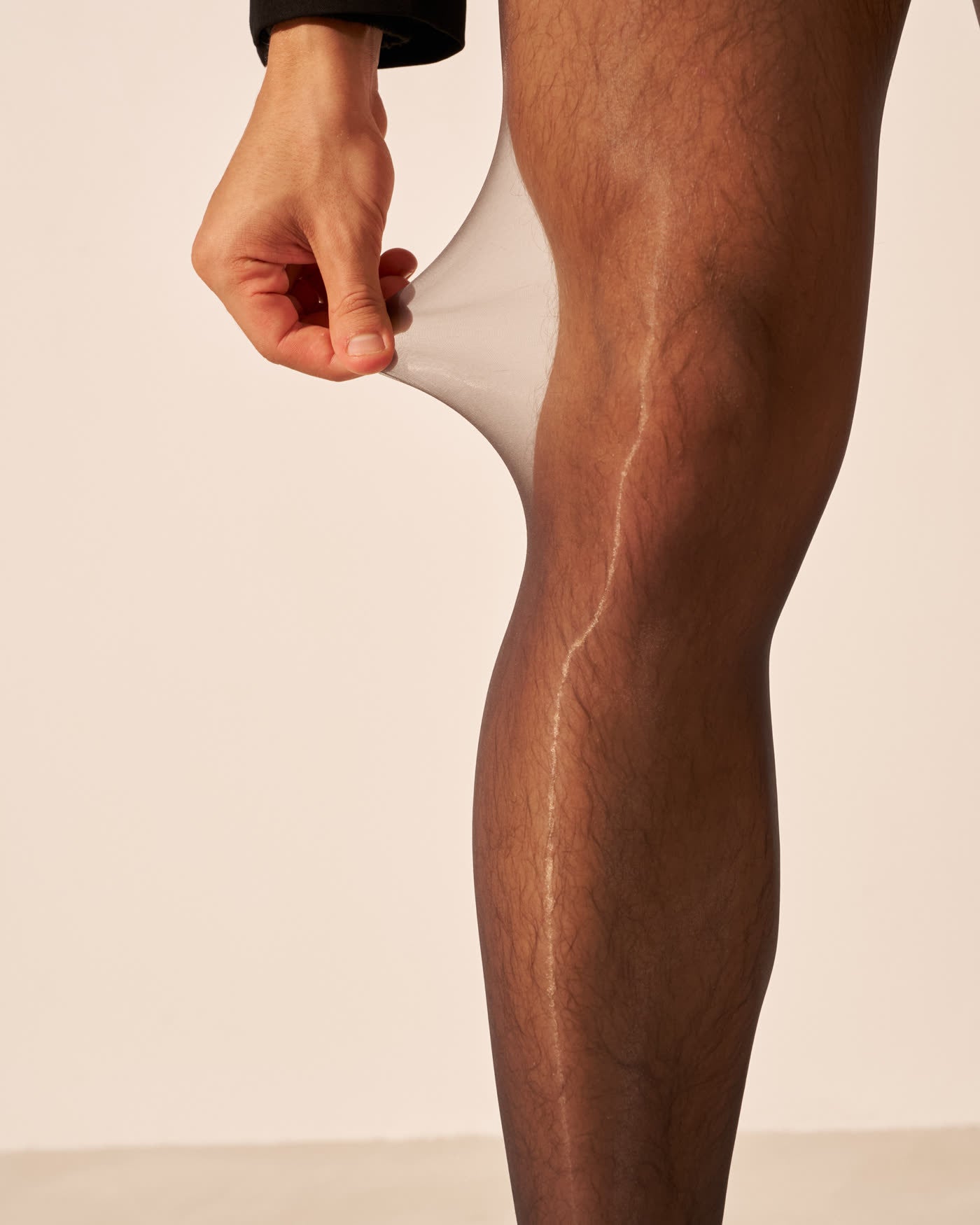 Close-up of fingers stretching sheer NYLO fabric away from the knee, showing the transparency of the knit
