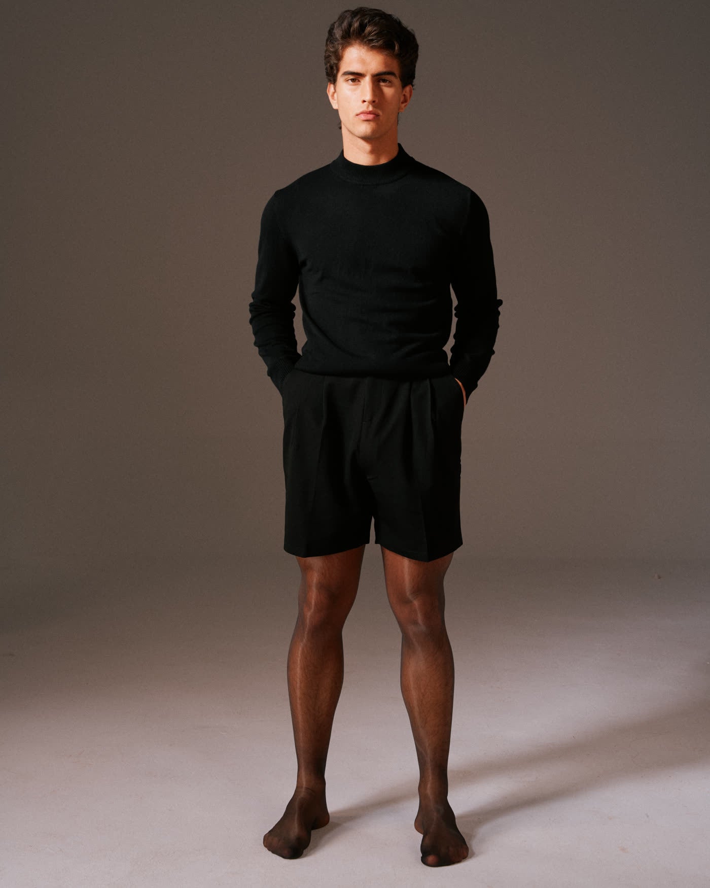 Man standing in a black mock neck and tailored shorts with sheer black NYLO tights, facing the camera